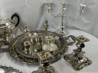 Full display of over 15 silver plated dining ware pieces on white table cloth, showing all items including candelabra, teapot, candlestick holders, cake stand, serving trays, and condiment holders.