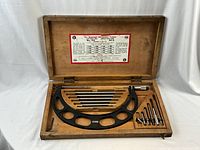 Open wooden case showing large black frame Starrett micrometer caliper with metal rods and extensions neatly arranged in slots.