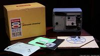 Photo showing the Kodak Brownie 8mm Movie projector Model A-15, its original yellow box with Brownie 8 branding, various documents, and a blue film take-up reel arranged on a surface.