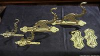 Photo showing all six brass decorative hardware pieces arranged on a dark fabric surface, including two gryphon motif curtain tie backs, two sea serpent motif rod holders, and two antique door push plates.