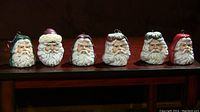 Six different porcelain Santa Claus head Christmas ornaments displayed in a row on a dark surface. Each ornament has distinct facial details and different hat styles, designed for hanging on a Christmas tree.