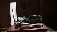 Photo showing black NuMe flat iron hair straightener, white branded container, black heat-resistant holder, and black protective carrying pouch with pattern and white stitching.