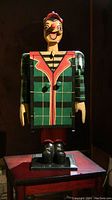 Full front body view of carved Pinocchio cabinet in green plaid jacket and red hat standing on a table.