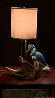 Folk art lamp with carved bird on wooden base, illuminated with light beige fabric shade and pull chain switch on a wooden surface against a dark background.