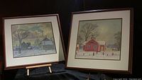 Two framed prints placed on a dark surface, one print shows a red schoolhouse and children playing outside in winter, the other shows a village winter scene with houses and snow.
