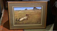 Framed print showing cat lying in grass with farm buildings in background, inside a brown wood frame and matte