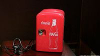 Front view of the mini Coca-Cola fridge with attached power cord coiled beside it, shows red body with white Coca-Cola logo and bottle graphic