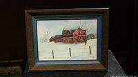 Framed folk art painting showing a red house and snow-covered landscape, displayed on a stand against a dark background.