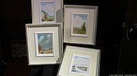 Four framed paintings displayed together, showing varied seasonal scenes each in white frames