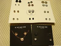 Overview photo of multiple pairs of stud earrings arranged on white sheet, and two Coach cards with jewelry set including necklace and earrings.