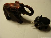 Photo showing two elephant figurines: larger reddish-brown wooden elephant with raised trunk and smaller dark wood elephant with painted tusks