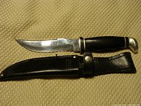 Whole hunting knife with blade extended alongside leather sheath on beige textured fabric background.