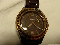 Front view of Fossil watch showing dark brown dial, gold tone bezel with amber rhinestones, and dark brown stainless steel band.