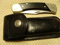 Folded Schrade LB7 pocket knife with visible monogram 'DJ' on silver bolster next to black leather sheath.