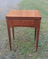 Front view of walnut sewing machine stand with single drawer and tapered legs outdoors on grass.