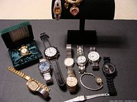 Full view of ten assorted watches and a desk clock displayed, showing various designs and bands including metal link, leather, and bracelet styles.