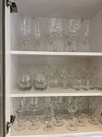 Three shelves of assorted clear glassware featuring different types of wine glasses, tumblers, and cocktail glasses inside a white cabinet.