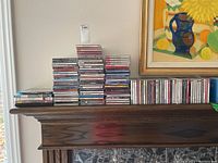 Full lot of approximately 60 CDs stacked on a wooden surface beneath a framed painting.