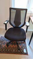 Front view of black swivel desk chair with mesh backrest and padded seat, armrests visible, placed on decorative rug near window.