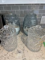 Four large glass storage containers on kitchen counter next to stove, two ribbed jars with black stripes and two clear crystal-like jars