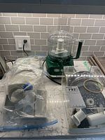 Green KitchenAid food processor with clear plastic bowl and lid, and various attachments in plastic bags and a tray