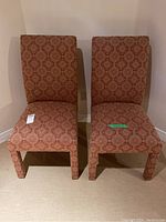 Two matching occasional chairs fully covered in patterned reddish fabric including legs, shown against a plain wall and carpeted floor.