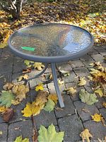 Round glass top patio side table on a stone or brick patio ground with fallen autumn leaves. Table has a gray metal frame and three legs with a textured clear glass surface.