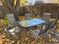 Front view of rectangular glass-top patio table surrounded by six metal sling chairs on a leaf-covered brick patio.