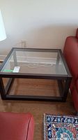 Full view of the square side table next to a red couch, showing wood frame and clear glass top.