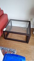Square side table made of dark wood frame with clear glass top on wood floor next to red couch.