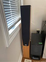 Front view of tower speaker with black grille installed, wood veneer cabinet beside window