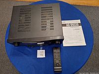 Top-down view of black TEAC AG-D9320 AV digital surround receiver with ventilation slots, remote control placed beside it, and the owner's manual laid out on blue surface.
