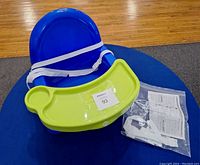 Blue plastic baby seat with a green detachable tray, white safety straps, and instruction manual on a blue round surface with wooden floor background.