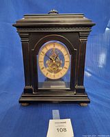 Front view of vintage black wooden desk clock with glass panels and visible inner gears behind the clock face with Roman numerals.
