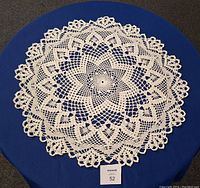 Top view of a large, white hand crocheted doily placed on a dark blue surface showcasing its detailed star and floral lace pattern and its approximate size of 28 inches diameter.