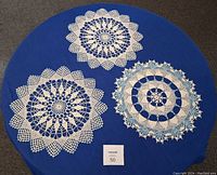 Photo showing three hand crocheted doilies displayed on a blue surface, with two mainly white doilies with floral patterns and one white and pale blue doily with circular patterns.