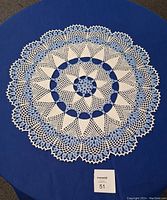 Top-down view of the large hand-crocheted doily laid flat on a blue surface, showing intricate blue and white crocheted circular patterns and measured at approximately 23 inches diameter.