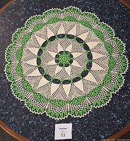 Top-down photo of a large round crocheted doily in green and ecru with a floral and geometric pattern on a dark surface.