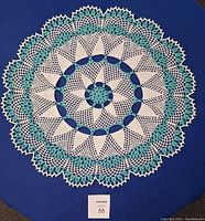 Top view of a large hand crocheted doily in blue and white yarn laid flat on a dark blue fabric background showing the intricate pattern.