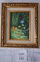 Photo of a small framed painting showing flowers, butterflies, and greenery, within an ornate gold-colored frame with decorative leaf motifs and linen-textured inner border.