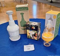 Three kitchen tools including a white Pampered Chef food chopper with original box, a citrus sprayer with orange base and box, and a Color Garden digital food scale in black box.