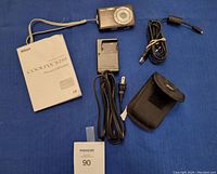 Photo showing Nikon Coolpix S210 digital camera with its instruction manual, carrying case, charger, cables, and battery arranged on blue cloth.