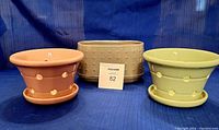 Three plant pots displayed on blue fabric backdrop: one beige oval pot with textured design, one pink round pot with holes and saucer, and one green round pot with holes and saucer.