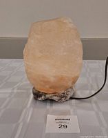 Photo of 8 inch tall salt crystal lamp on a table, showing its natural rough crystal shape on a salt base with attached black electrical cord.