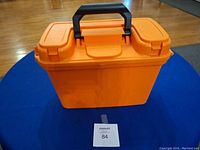 Closed orange plastic tool box with black handle on top and two latch closures, sitting on a blue surface with visible wood floor background.