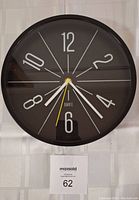 Front-facing image of the round black wall clock with white markings and hands on a light-colored wall.