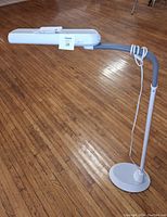White tall bendable work lamp standing on wooden floor, showing adjustable neck, round base and rectangular lamp head