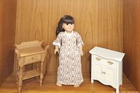 Full view of the Samantha doll flanked by the 1993 wooden nightstand on the left and the white washstand/dresser on the right against a wooden backdrop.