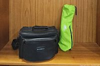 Black Ambico branded carrying case alongside green tripod bag on wooden background.