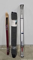 Three items against white wall and floor: boxed shower rod and curtain rod standing upright, and rolled floor mat in center wrapped in plastic.